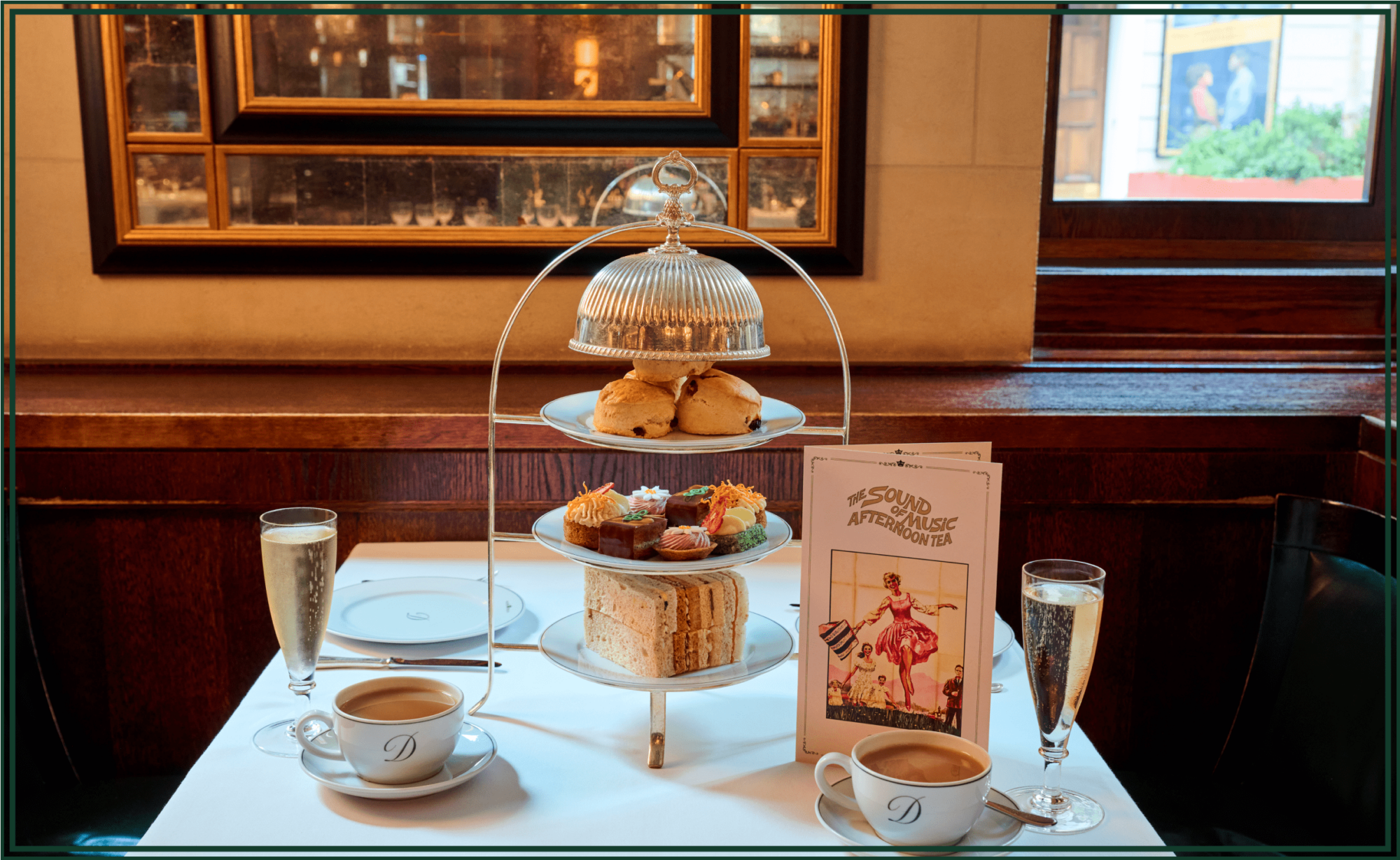 A beautifully set afternoon tea table for two at a vintage-style venue. The table features a three-tiered silver stand holding scones on the top tier, colorful pastries and cakes in the middle, and finger sandwiches on the bottom. Two cups of tea in white cups marked with a "D" sit on either side of the stand, each accompanied by a flute of sparkling wine. A themed menu titled "The Sound of Music Afternoon Tea" with a vintage-style illustration is placed on the table. The setting is cozy, with wooden paneling, a window view, and a mirrored wall in the background.